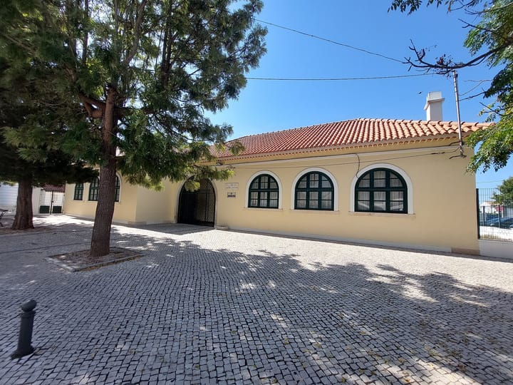 A single-storey yellow school canteen building with arched green windows and a red tiled roof beside a cobbled square in Salvaterra de Magos