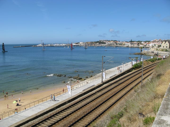 The bay of Cascais seen from above the coastal railway at Estoril, with a sandy beach, the seafront promenade, the marina and the white town beyond
