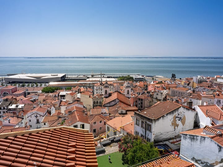 The red-tiled rooftops of Alfama running down to the Tagus in Lisbon, seen from the Miradouro de Santa Luzia on a clear day, with the cruise terminal on the waterfront