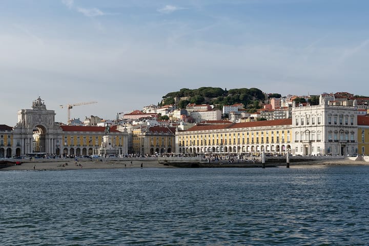 Lisbon's Praça do Comércio seen from the Tagus, its yellow arcades and triumphal arch lined with visitors, and the castle hill rising behind