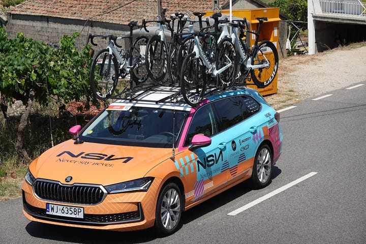 A cycling team car with racing bikes on its roof rack driving past vines and a stone house during the 2026 Volta a Portugal in Melgaço