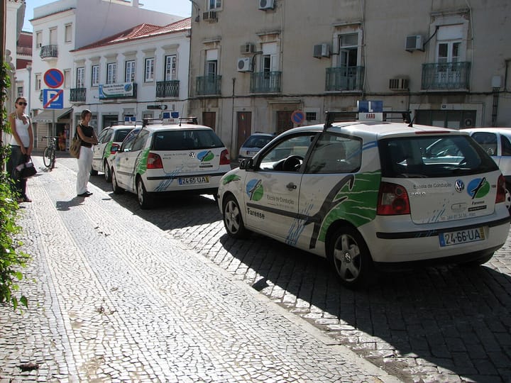 White driving school cars with L plates on the roof parked on a cobbled street in Faro, as learner drivers wait on the pavement