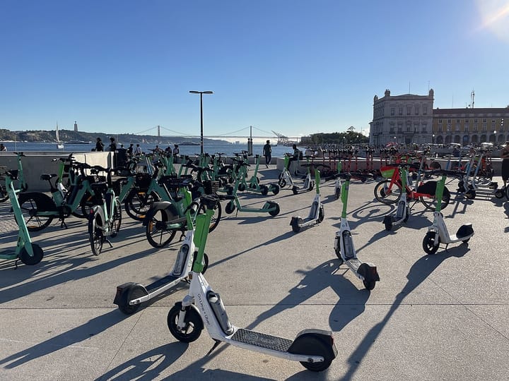 Rows of shared electric scooters and bikes parked on the riverside at Praça do Comércio in Lisbon, with the 25 de Abril bridge in the distance
