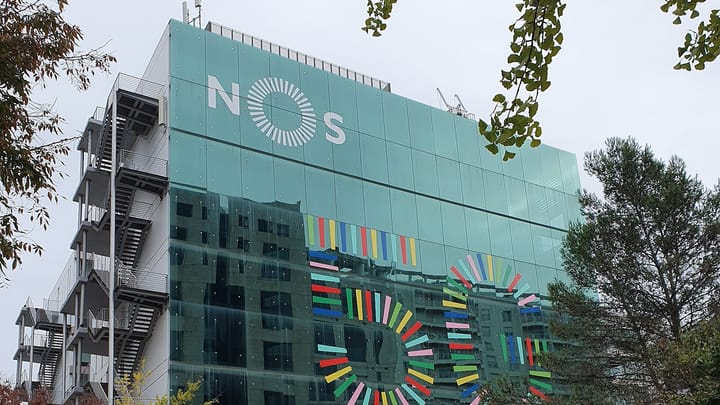 The NOS headquarters in Lisbon, a green glass block carrying the NOS wordmark near its roofline above a large coloured mural, with an external steel stair on the left