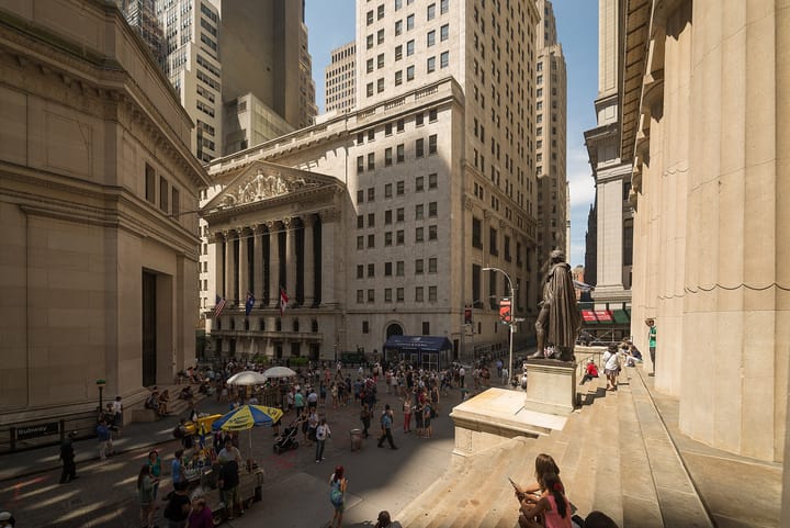 The columned facade of the New York Stock Exchange on Broad Street, seen from the steps of Federal Hall, with crowds in the plaza and the George Washington statue at right