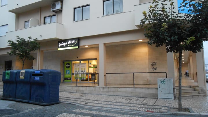 A lit Pingo Doce supermarket sign above a shop entrance at dusk in Figueira da Foz, with recycling bins and calcada pavement in the foreground