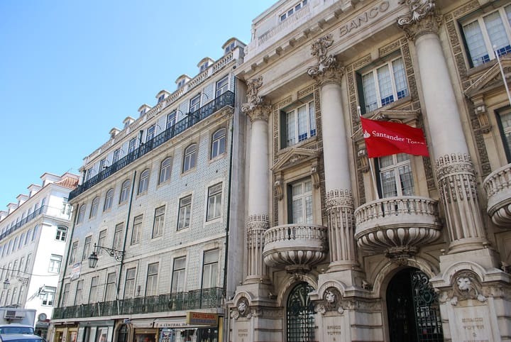 The columned stone facade of the Banco Totta e Acores building on Rua do Ouro in Lisbon, flying a red Santander Totta flag, with a tiled townhouse beside it