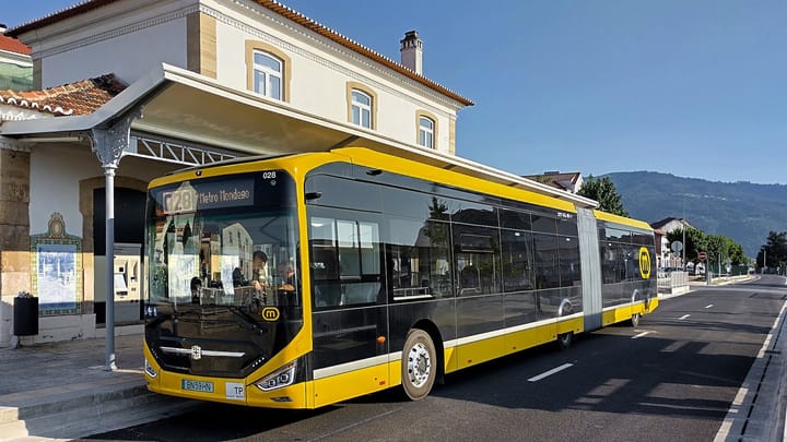 A yellow and black articulated electric Metrobus of Metro Mondego stops at the restored Lousã station building under a clear blue sky, with hills behind