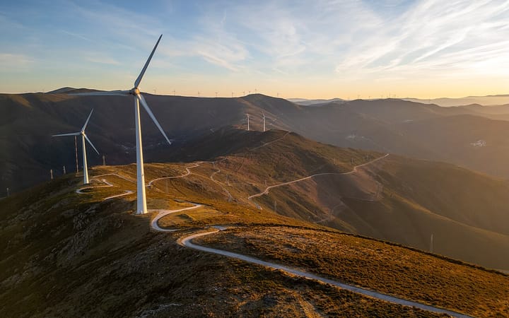Wind turbines along a mountain ridge at the Colcurinho wind farm in central Portugal, with a gravel service road winding below at sunset