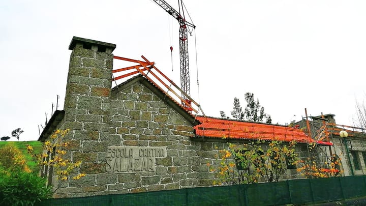 The granite Escola Cantina Salazar in Vimieiro, its name carved on the gable wall, during roof restoration work with a crane and new orange tiles