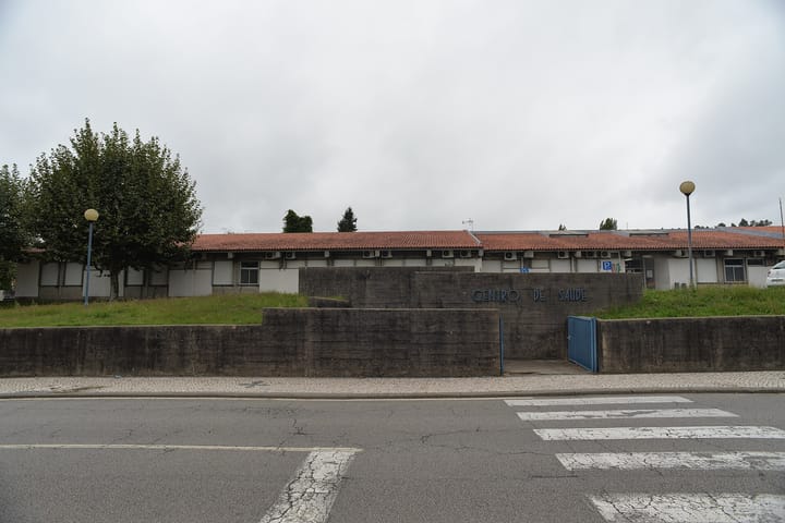 A low health centre building with a terracotta roof behind a concrete wall marked Centro de Saúde in Vale de Cambra