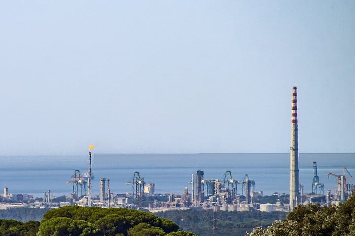 The Sines industrial complex seen across pine woods, with a tall striped chimney, a gas flare and port cranes against the sea